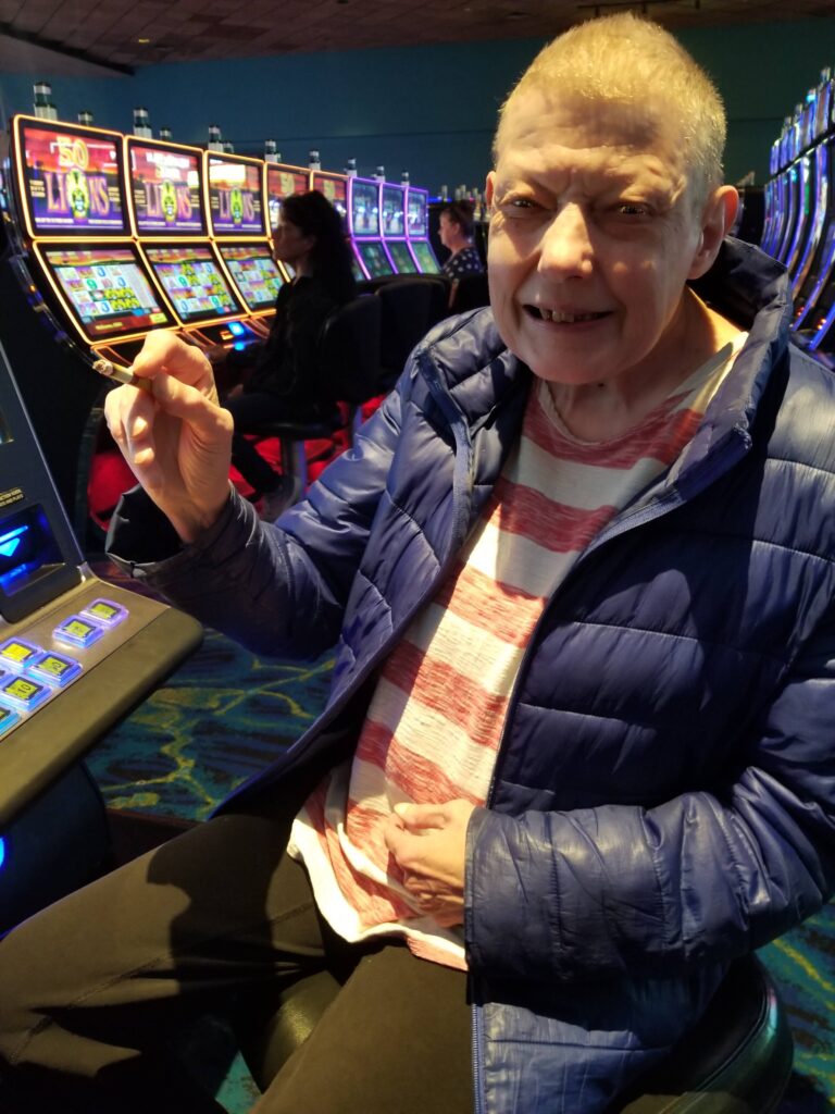 A woman is sitting at a slot machine in a casino. She is smiling and smoking a cigarette.