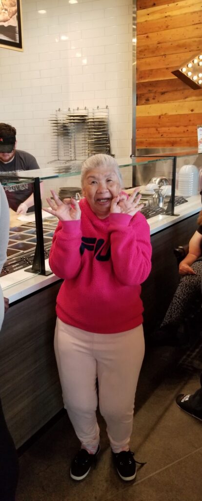 A Vietnamese woman with grayish-white hair is standing in front of the countertop at Mod Pizza. She is smiling and making the "OK" sign with both of her hands held up.
