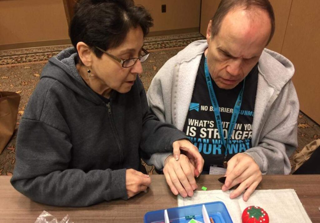 A woman is sitting next to a man who appears to be blind. She places her hand over his and guides his hands over the craft supplies.