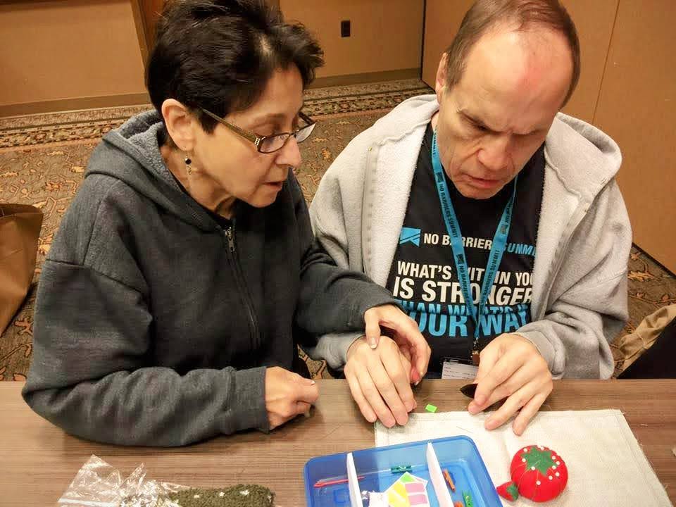 A woman is sitting next to a man who appears to be blind. She places her hand over his and guides his hands over the craft supplies.