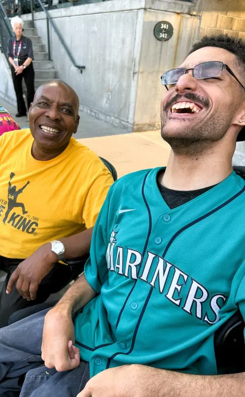 Two men are seated in the stadium at a Mariner's baseball game.